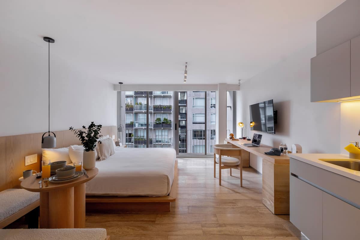 A modern hotel room with a bed, bedside table, desk, and a view of the cityscape through large windows.