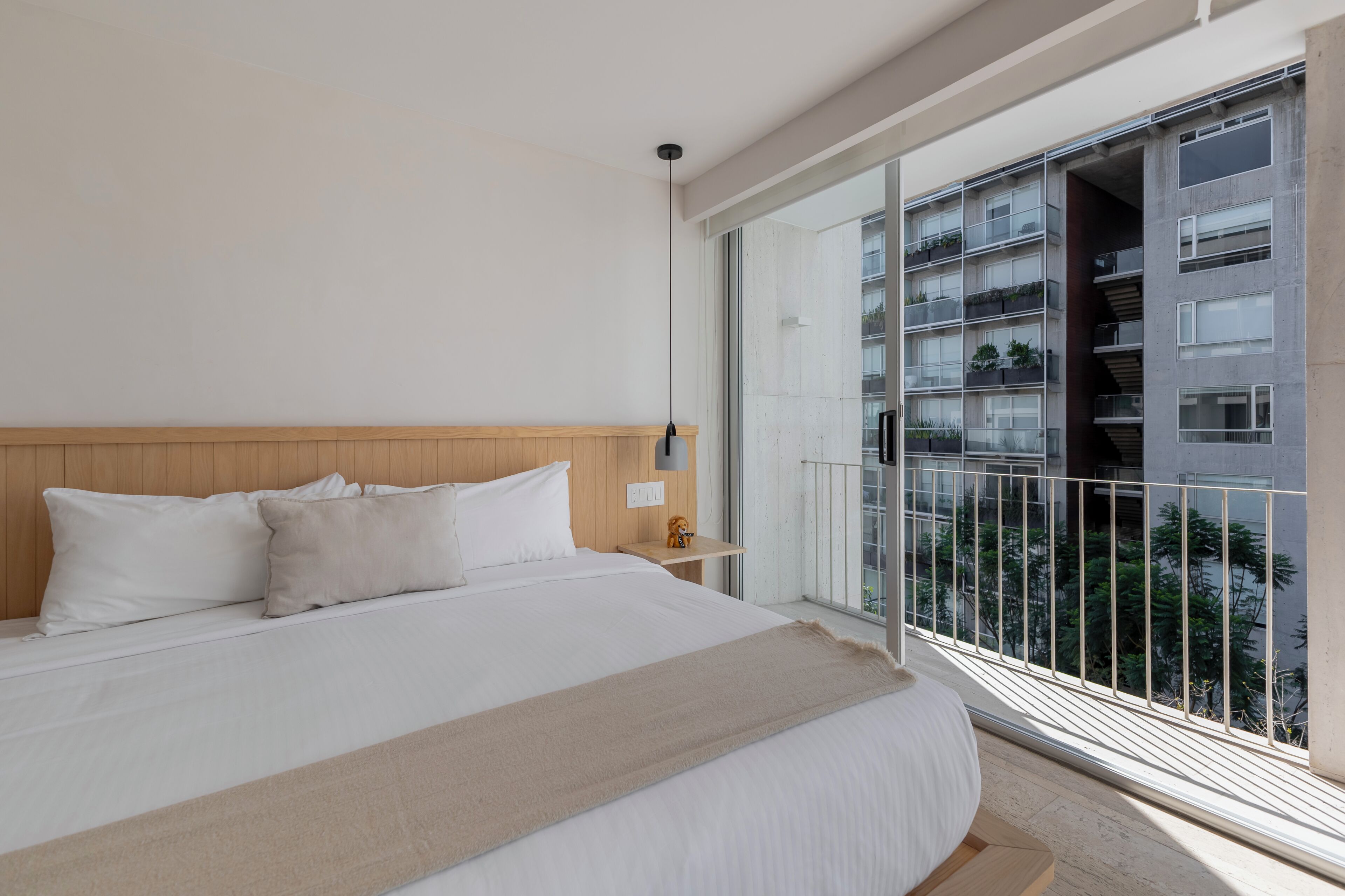 Un dormitorio con una cama grande, una mesita de noche, una lámpara de pared y un balcón con vistas a otros edificios.