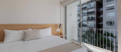 Un dormitorio con una cama grande, una mesita de noche, una lámpara de pared y un balcón con vistas a otros edificios.