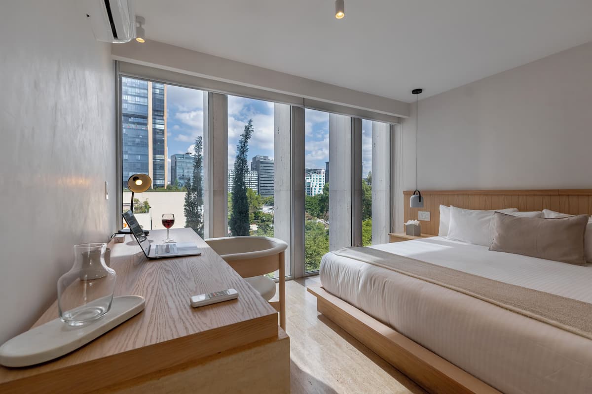 A modern hotel room with a large window, a bed, a desk with a laptop, and a remote control.
