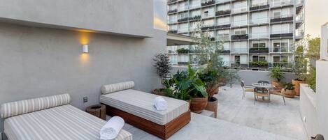 A modern outdoor seating area with two cushioned benches and a small table, surrounded by potted plants and a high-rise building in the background.