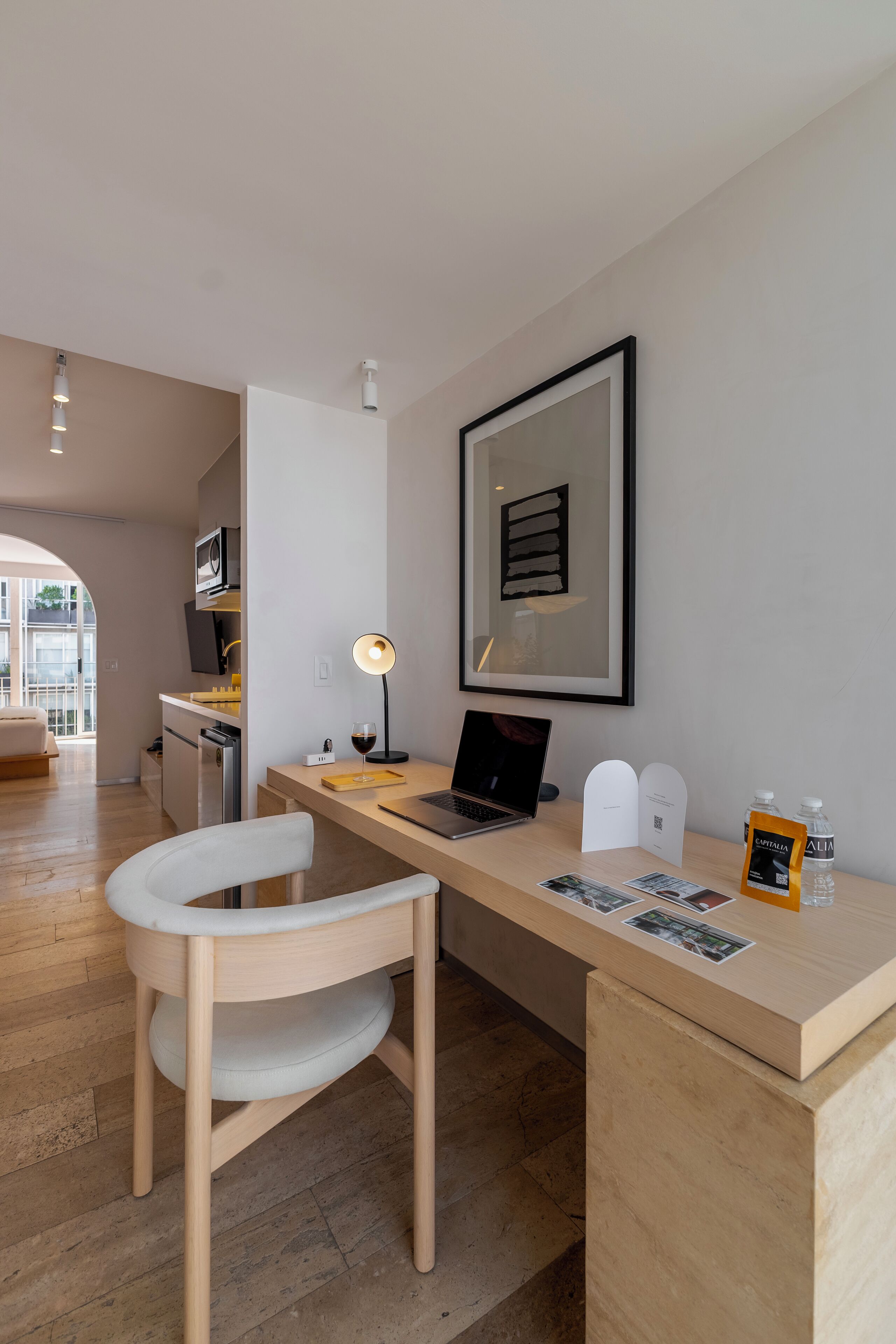 Une pièce moderne et bien éclairée, avec un bureau en bois, un ordinateur portable, une chaise, un tableau encadré au mur et une petite table sur laquelle se trouvent une bouteille d’eau et une photographie encadrée.