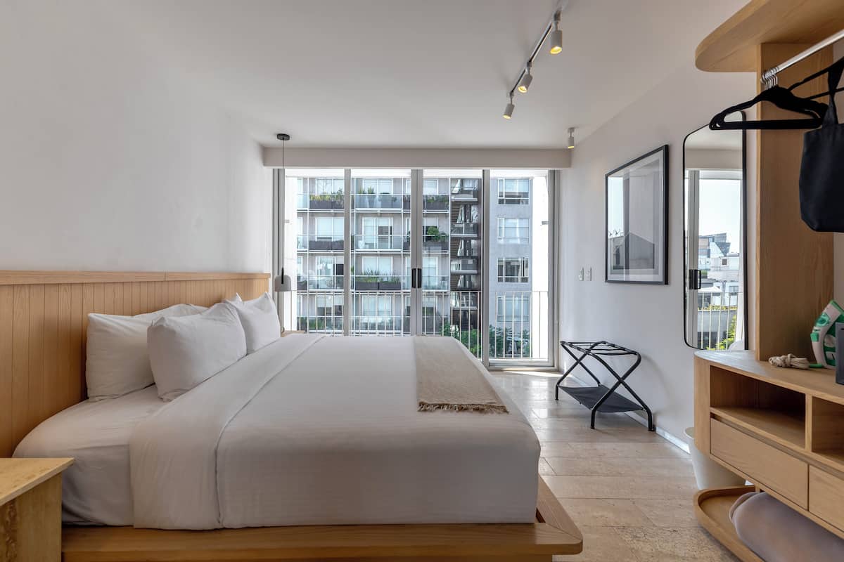 A modern hotel room with a large bed, wooden headboard, a wooden bench, a small table, a hanging chair, and a large window with a view of a building and balconies.