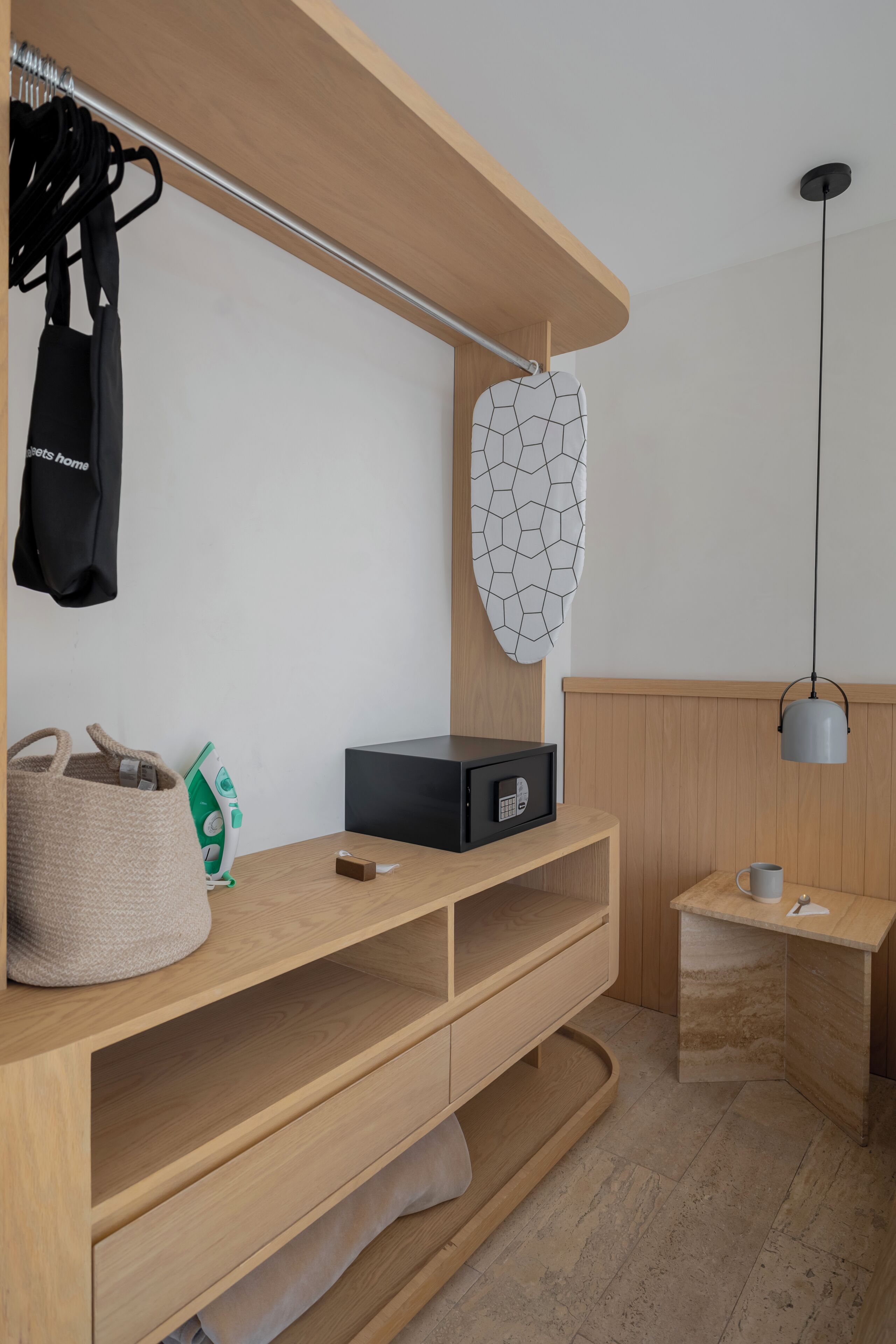 Une chambre moderne avec un bureau en bois, un sac suspendu, une planche à repasser et un élément décoratif fixé au mur.