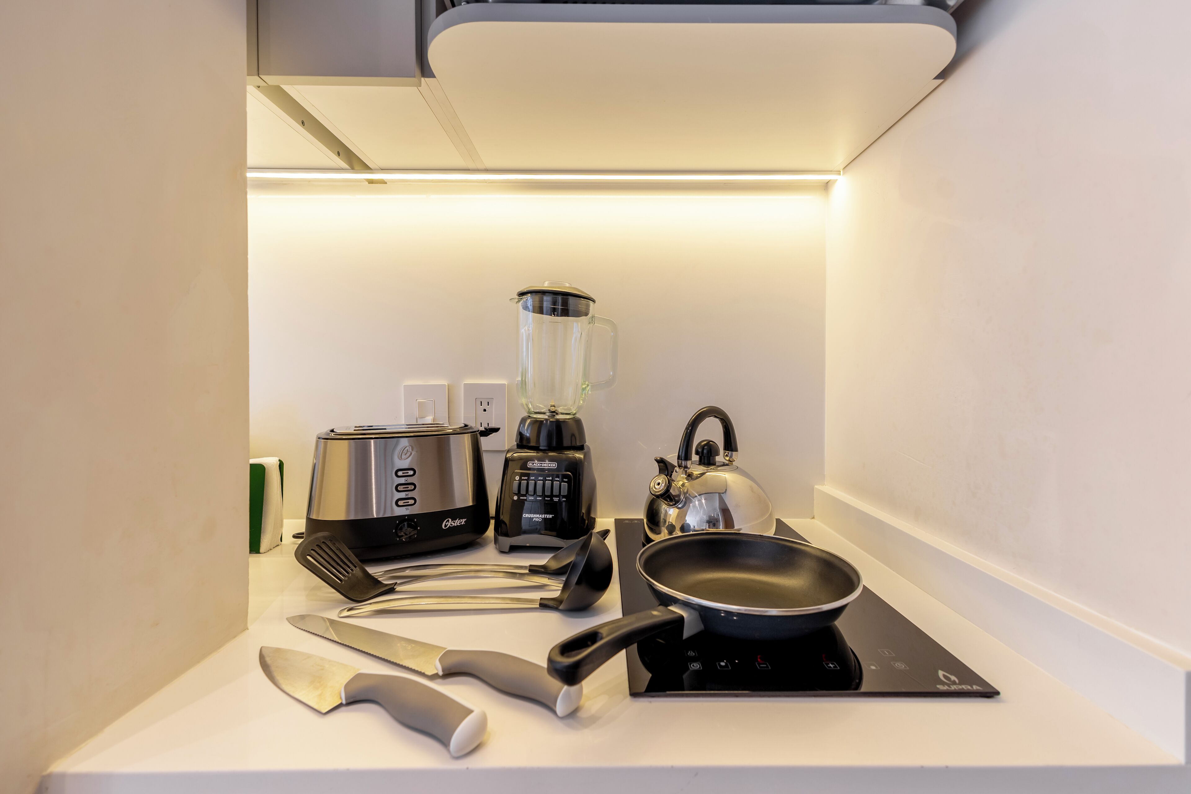 Een compact keukenhoekje met een broodrooster, blender, waterkoker, koekenpan en bestek op een wit aanrecht.