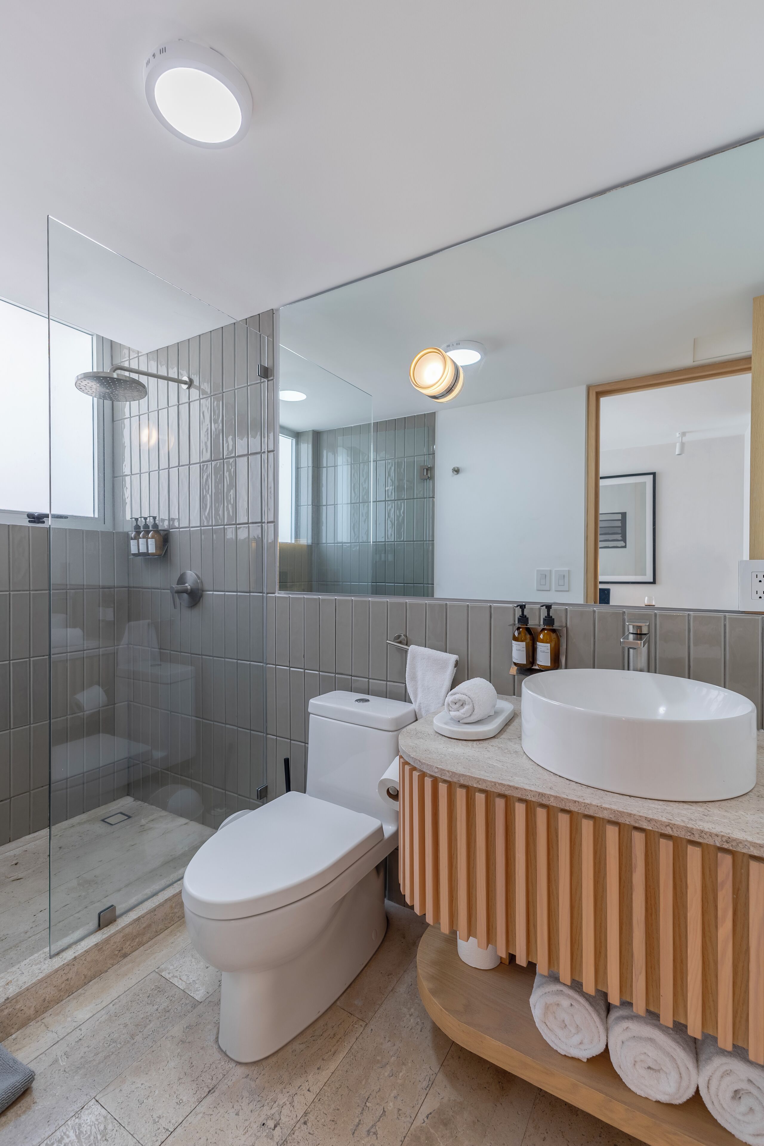 Un baño moderno con ducha de vidrio, inodoro blanco, un lavamanos grande de color blanco y un mueble de tocador de madera con toallas.
