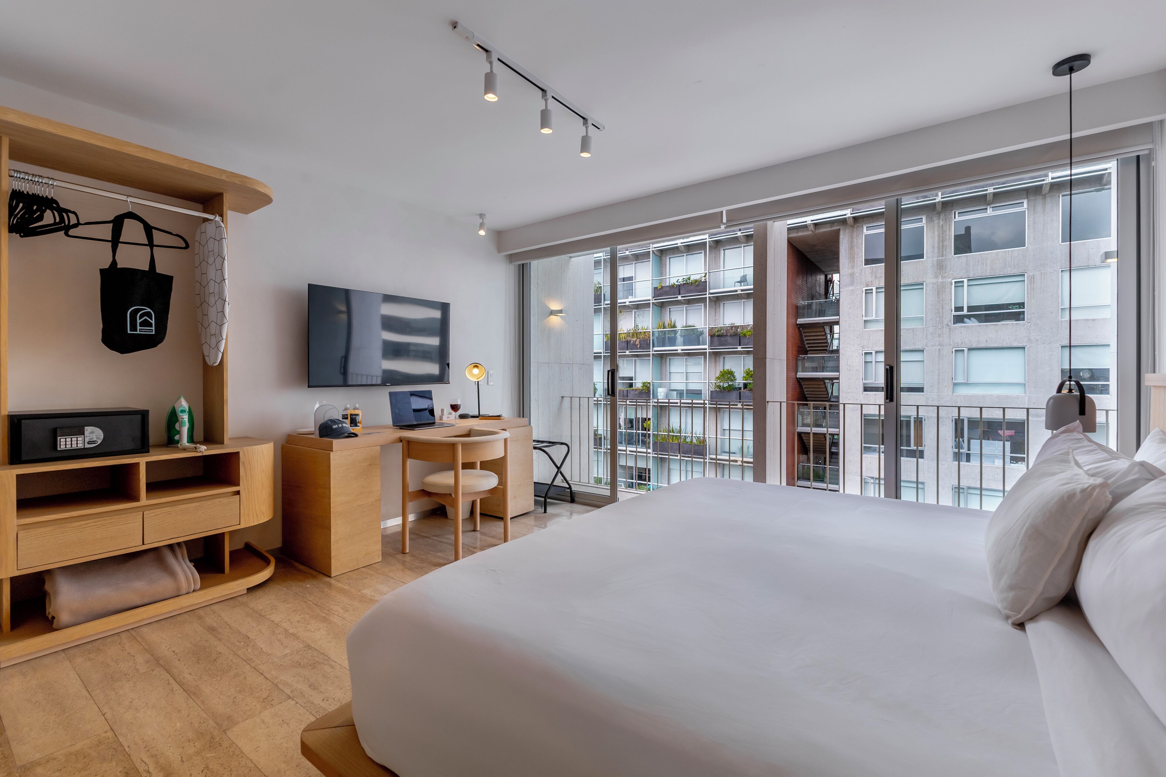 A modern hotel room with a large bed, a desk with a laptop, a chair, a wardrobe, and a balcony with a view of the city.