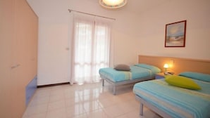 A bedroom with two beds, a window with curtains, a bedside table with a lamp, and a painting on the wall.