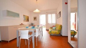 A modern kitchen with white cabinets, a dining table with chairs, and a colorful sofa in a bright living area with orange flooring.