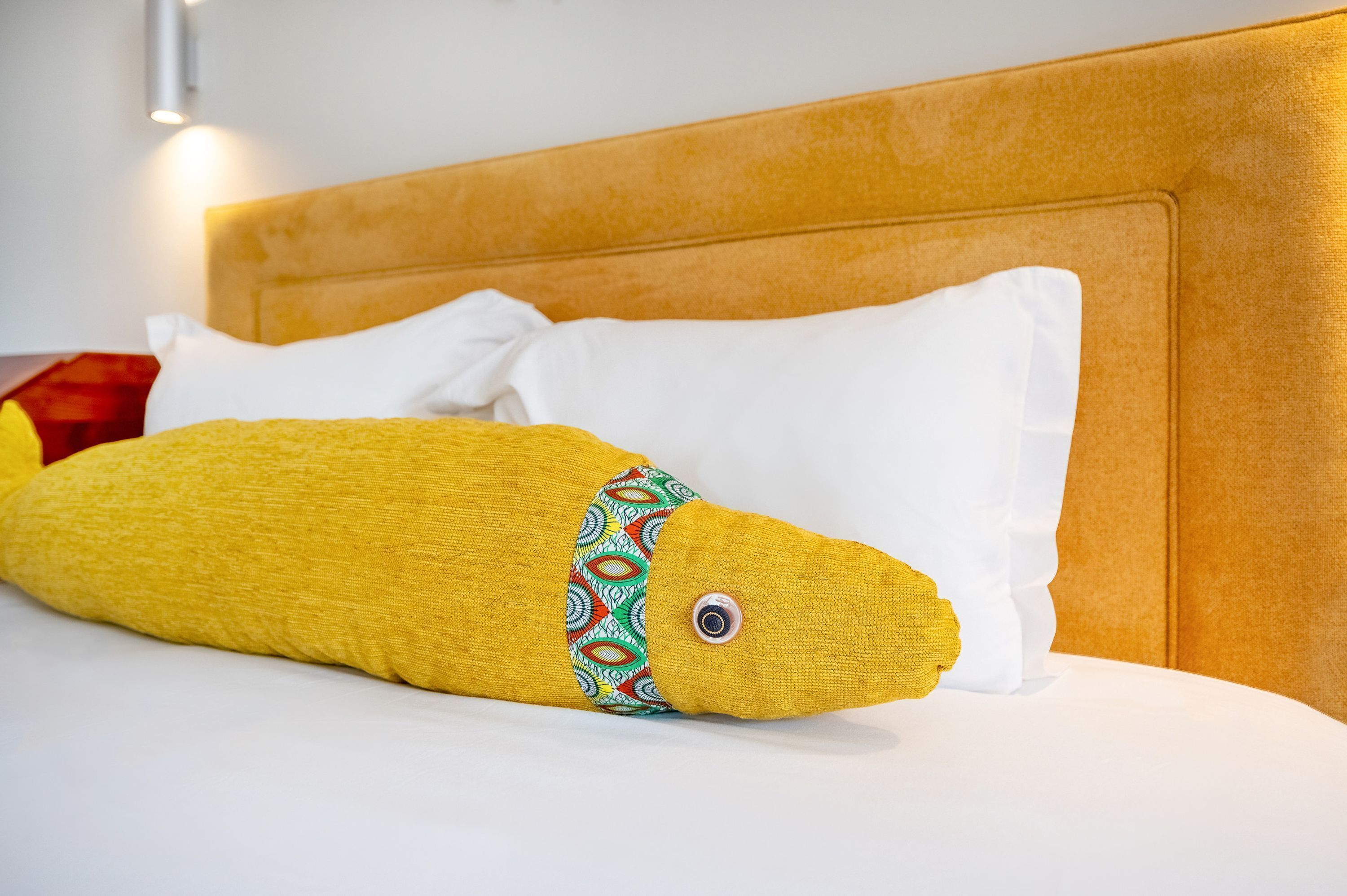 Een bed met een geel kussen en witte kussens, een houten hoofdbord en een rood object op de achtergrond.