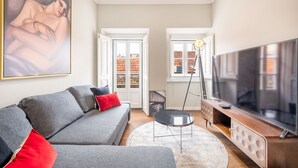 A modern living room with a grey sofa, a red pillow, a black coffee table, and a large flat-screen TV on a wooden stand. - Bairro Alto Prime Duplex by Homing (Lisbon)