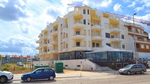 Un edificio de varios pisos con balcones, una entrada visible y un estacionamiento con coches.
