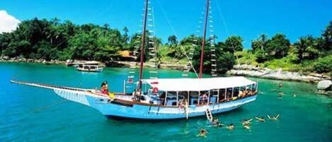 A boat with multiple decks and passengers, surrounded by clear turquoise water and a tropical landscape with trees and rocks.