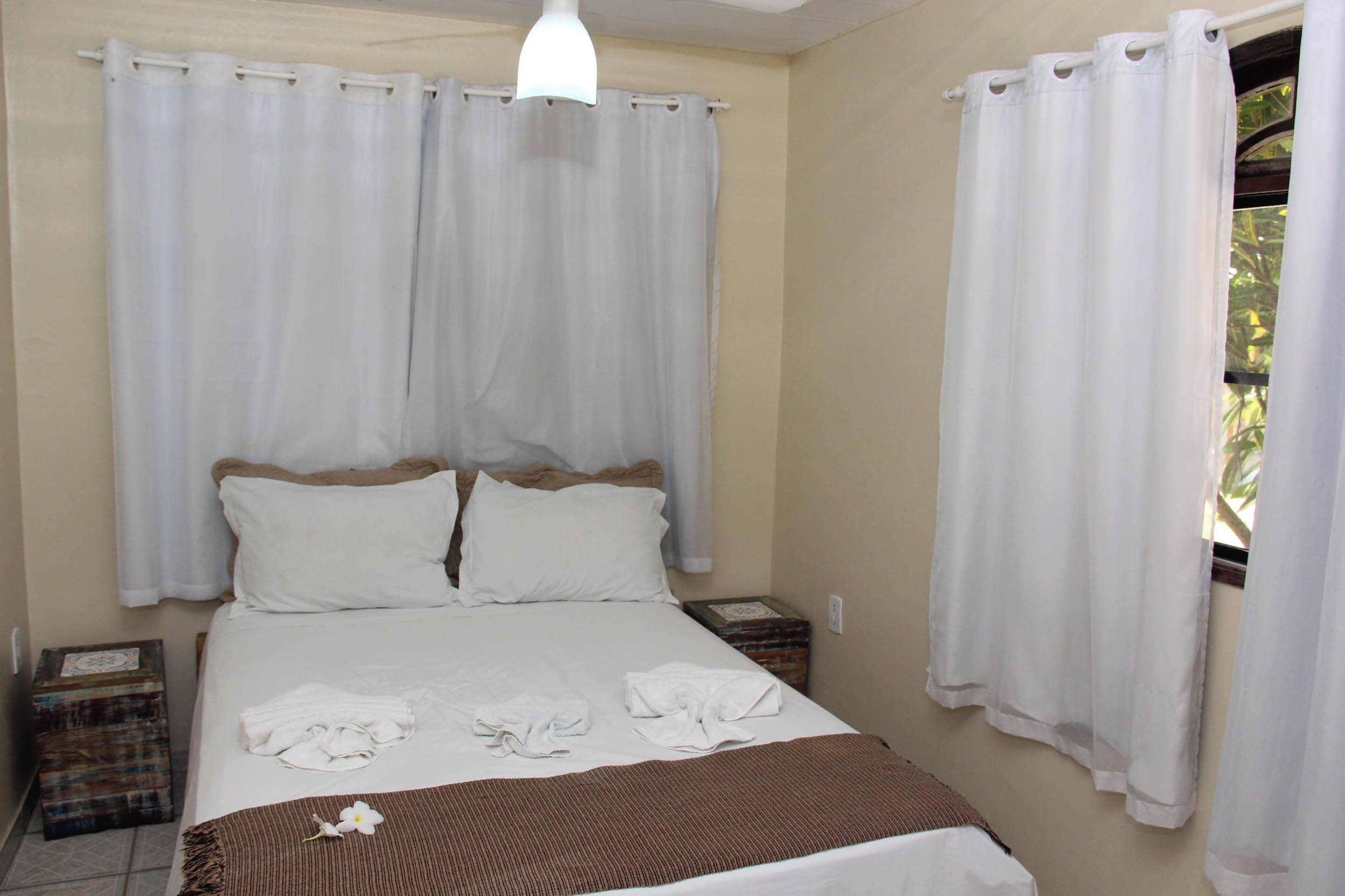 Un dormitorio con cama, cortinas blancas, una ventana con vistas y una mesita de noche.