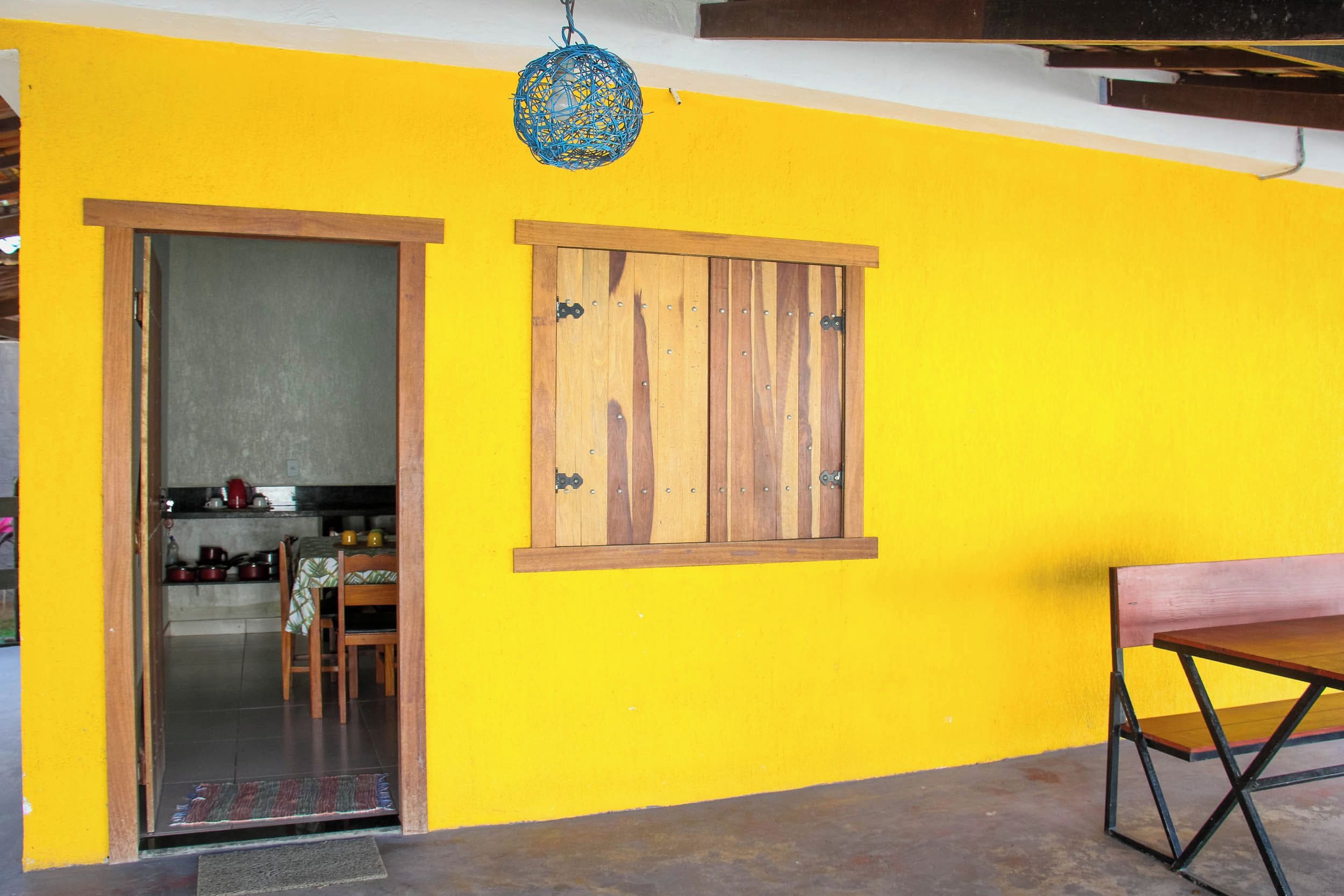 A room with a yellow wall, a wooden door, a window with wooden shutters, a dining table, and a hanging light fixture.