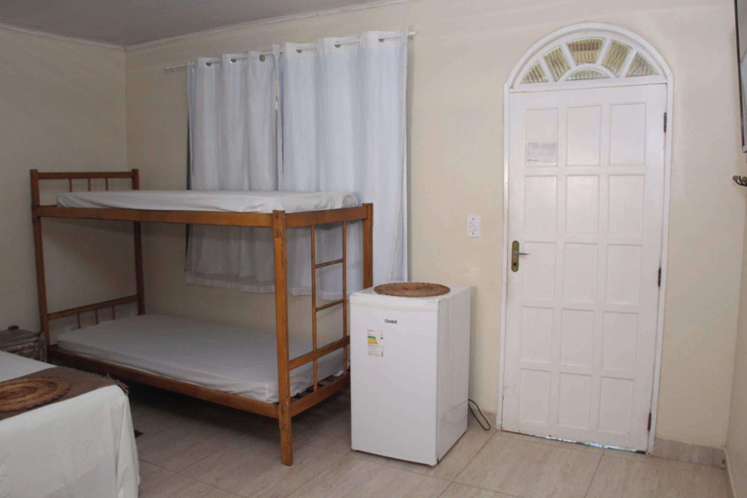 Une chambre avec un lit superposé, un petit réfrigérateur et une porte blanche surmontée d’une fenêtre.