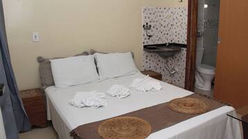 Habitación de hotel con cama, mesita de noche, lavamanos pequeño y un inodoro visible a través de una puerta abierta.