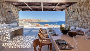 An outdoor dining area with stone walls, a view of the sea, and a long table set for dining. - Villa in Mykonos with 10 bedrooms sleeps 20 (Mykonos)