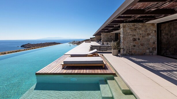 An infinity pool with a wooden deck and lounge chairs, leading to a stone building with a view of the sea. - Villa in Mykonos with 10 bedrooms sleeps 20 (Mykonos)