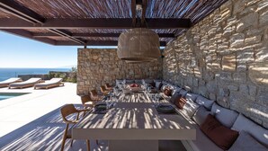 An outdoor dining area with stone walls, a long table set for dining, and lounge chairs by a pool. - Villa in Mykonos with 5 bedrooms sleeps 10 (Mykonos)