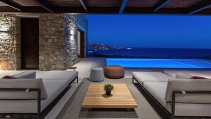 A modern outdoor seating area with stone walls, a wooden table, and a view of the sea at night. - Villa in Mykonos with 5 bedrooms sleeps 10 (Mykonos)