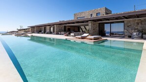 Una vila moderna amb una gran piscina infinita, façana de pedra i zona d'estar a l'aire lliure.