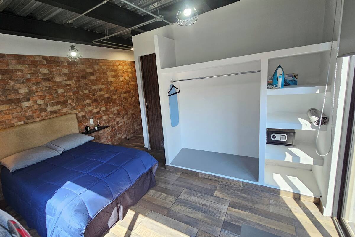 Un dormitorio con pared de ladrillo, una cama con colcha azul, una mesita de noche, una lámpara colgante y un armario con espejo y estantes.