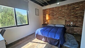 Een slaapkamer met een groot raam, een bakstenen muur, een bed met blauw beddengoed, een bedlampje en een klein plankje met een klok en een plant.