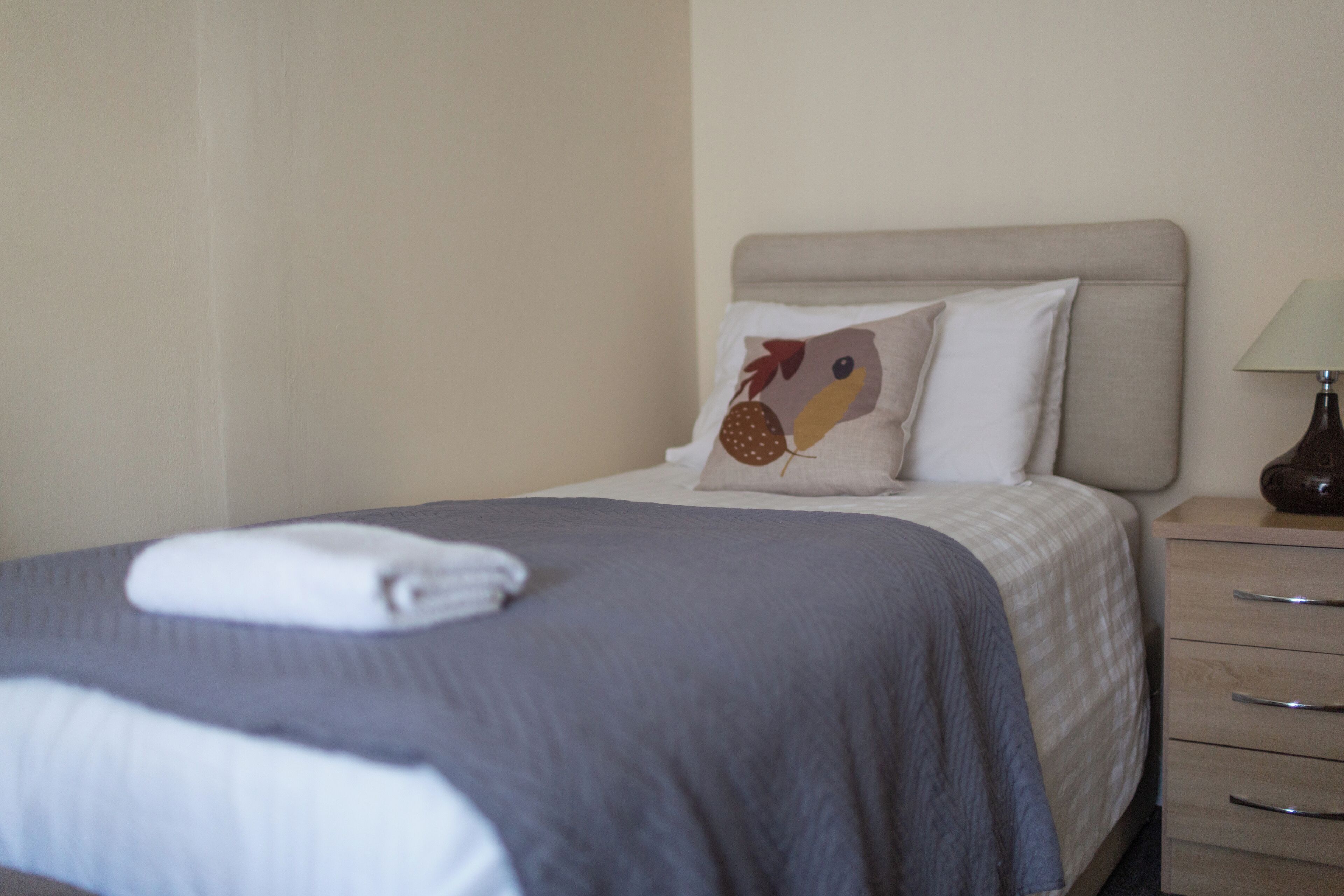 A neatly made bed with a grey bedspread and two pillows, one with a bird design.