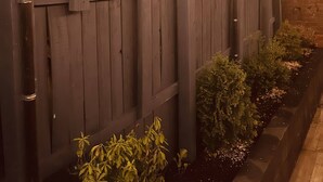 Una cerca de madera con paneles decorativos, un muro de ladrillo y un cantero con plantas.