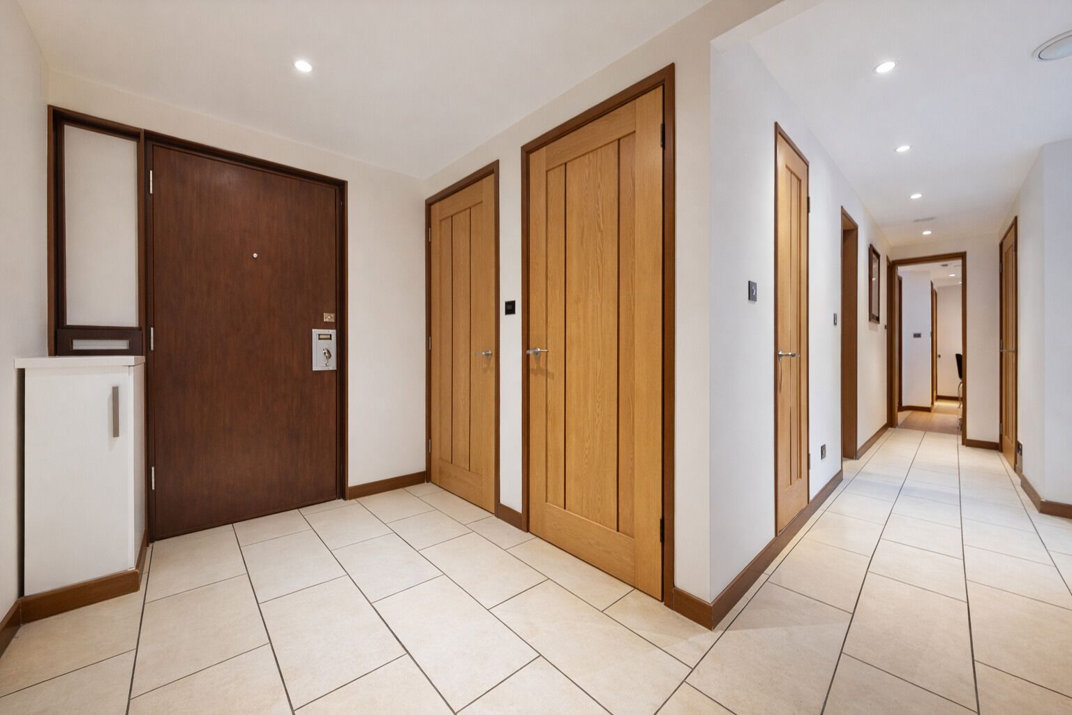 Un pasillo de hotel con puertas de madera, un refrigerador blanco y suelo de baldosas.