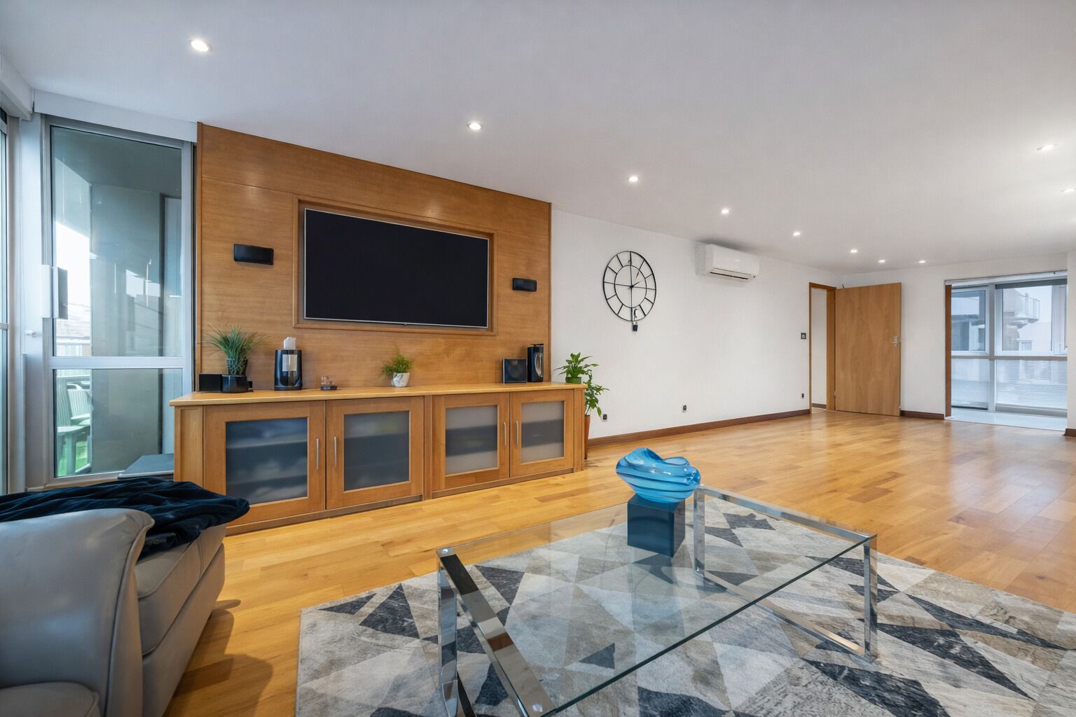 A modern living room with a wooden cabinet, a flat-screen TV, a clock, and a glass coffee table.