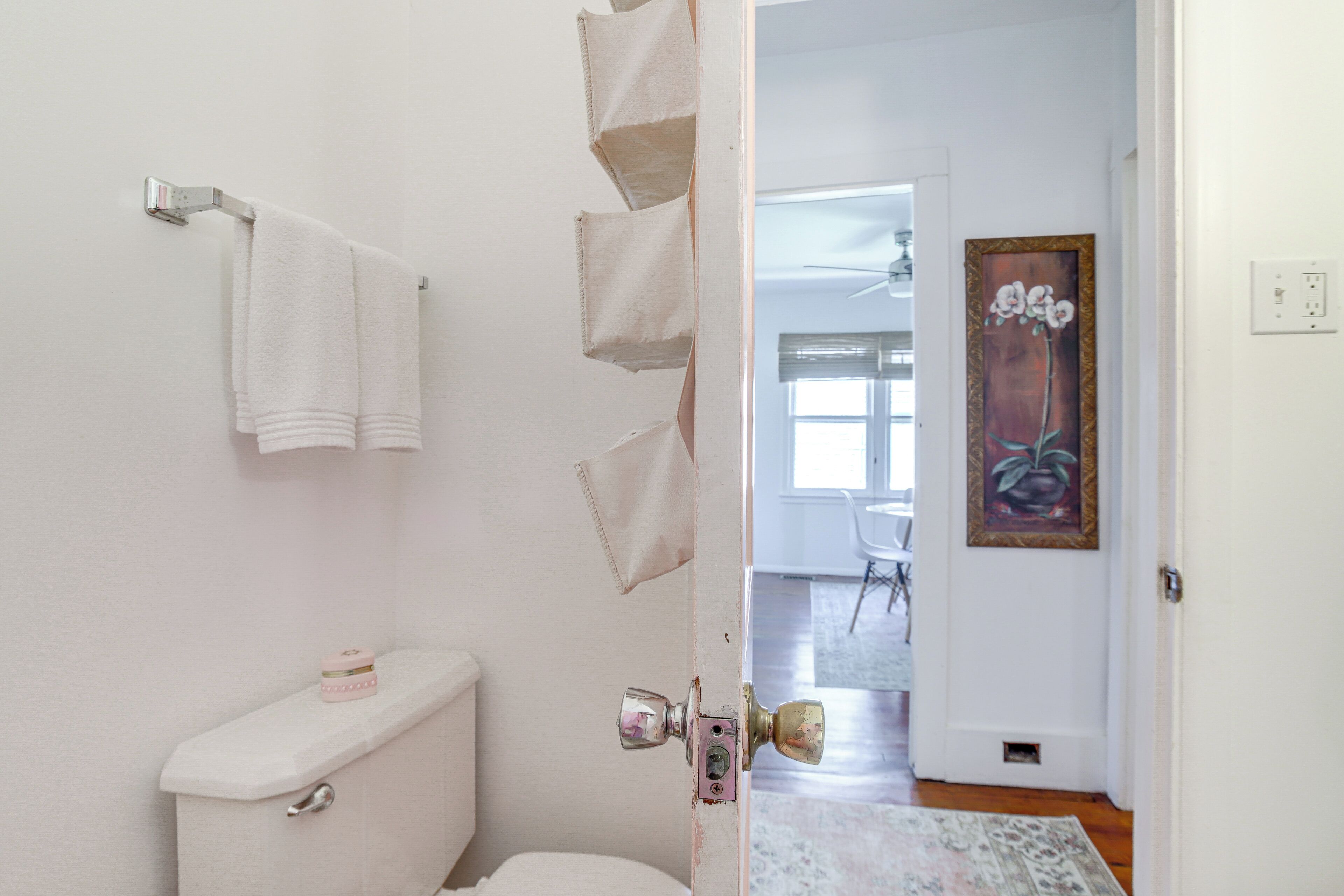A bathroom with a toilet, a towel rack, and a door leading to a living area with a rug and a painting.