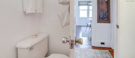 A bathroom with a toilet, a towel rack, and a door leading to a living area with a rug and a painting.