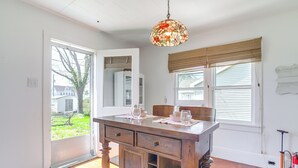 Een eethoek met een houten tafel, stoelen en een hanglamp van geëtst glas. Er is een raam met zonwering en uitzicht op buiten.
