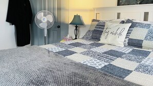 A bedroom with a bed, a standing fan, a door, a lamp, and a painting on the wall.