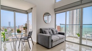 A modern living room with a grey sofa, a round mirror, and a glass table with chairs. Large windows offer a view of the city and the sea. - Home Beach MagnoLoft (Cartagena)