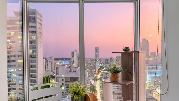A modern balcony with a glass table and chairs, surrounded by potted plants and a wooden shelf. - Home Beach MagnoLoft (Cartagena)