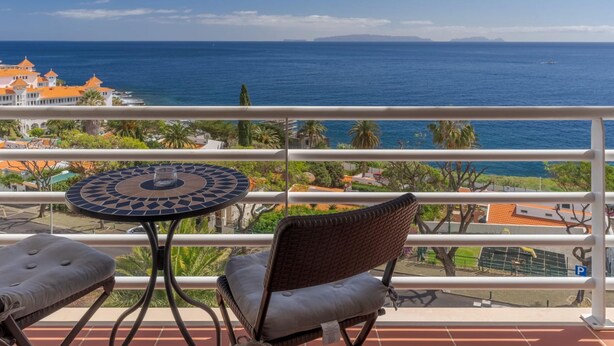 A balcony with a table and chairs, overlooking a coastal view with buildings and palm trees. - Apartamento Estrela Azul (Santa Cruz)