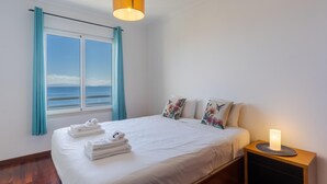 A bedroom with a large bed, a bedside table with a candle, and a view of the sea through a window with blue curtains. - Apartamento Estrela Azul (Santa Cruz)