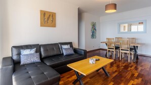 A modern living room with a black leather sofa, a wooden coffee table, and a dining area with wooden chairs and a table. - Apartamento Estrela Azul (Santa Cruz)