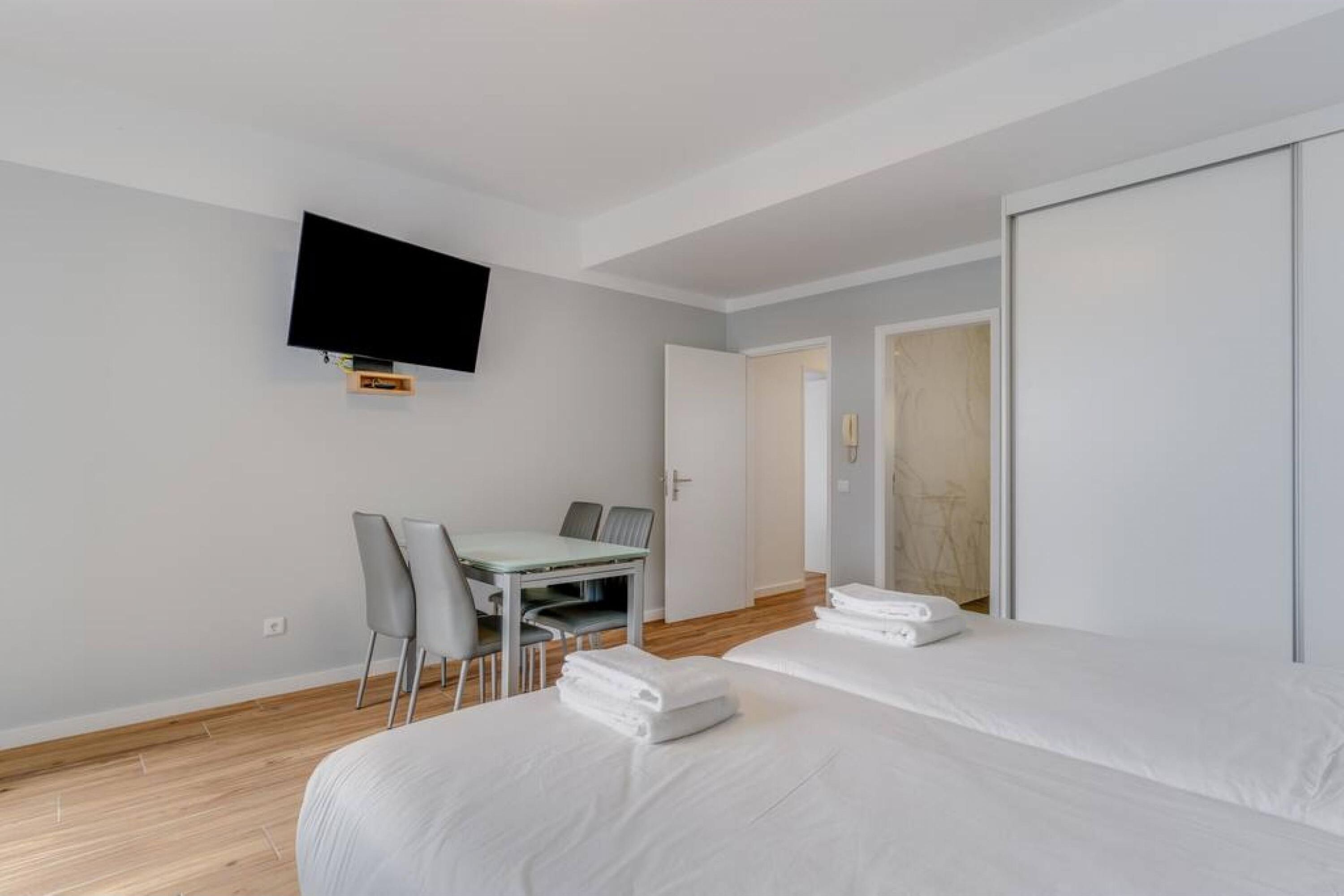 A hotel room with two beds, a TV mounted on the wall, a dining table with chairs, and a door leading to another room.