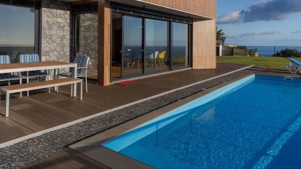 A modern poolside area with a wooden deck, a pool, and a building with large glass doors. - Casa Mozart II 15B in Estreito da Calheta (Calheta)