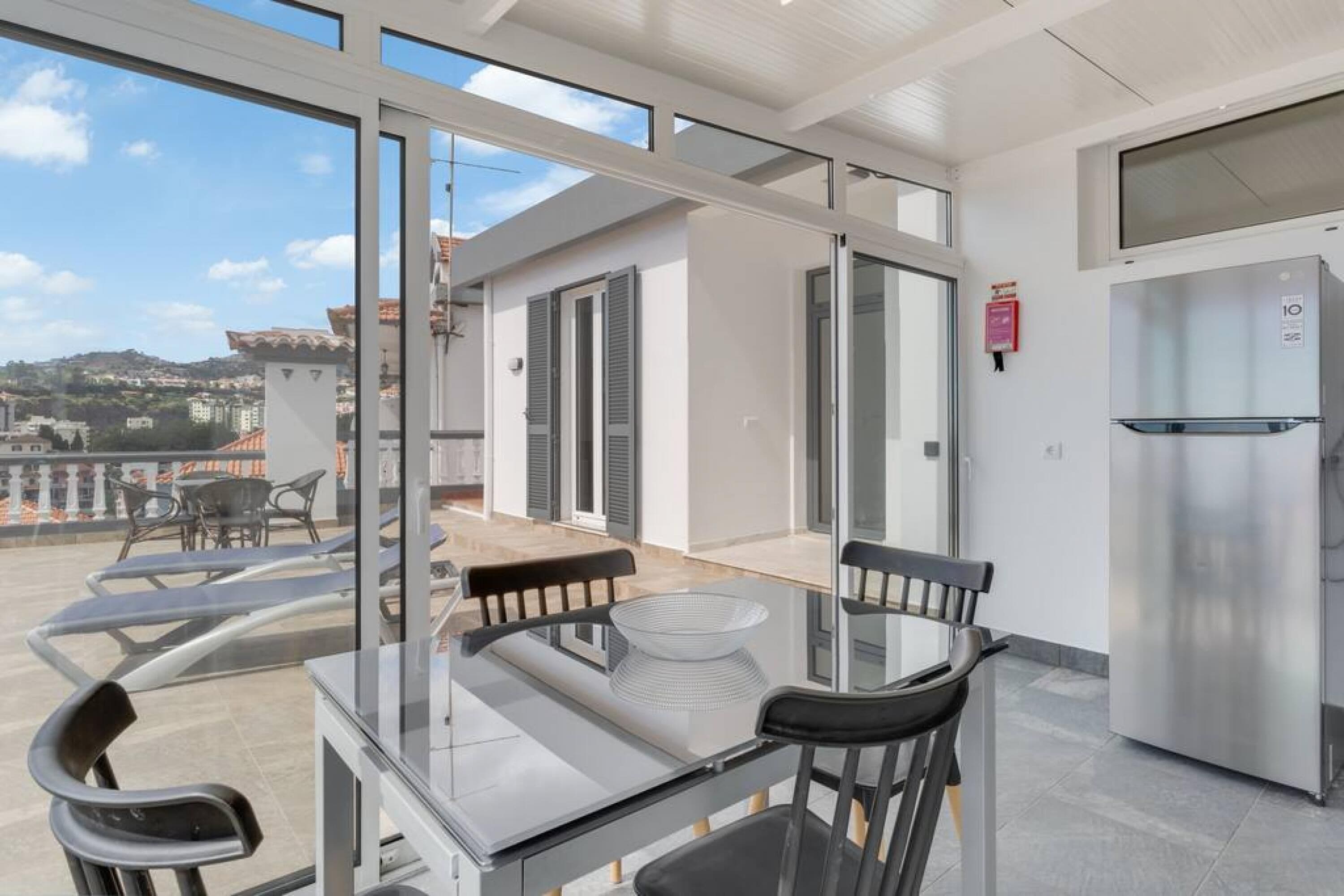 Une cuisine moderne dotée d’un réfrigérateur en acier inoxydable, d’une table à manger en verre avec des chaises, et offrant une vue sur la ville grâce à de grandes fenêtres.