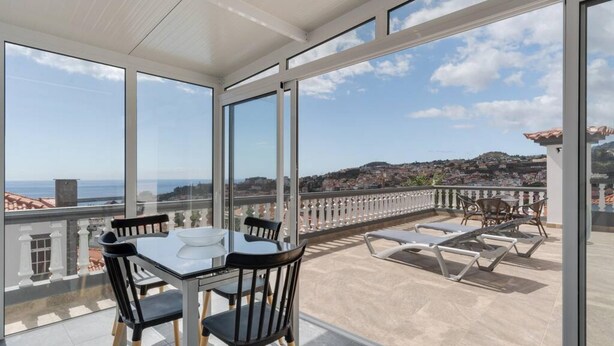 En balkon med et bord og stole, havudsigt og en bysilhuet i baggrunden.