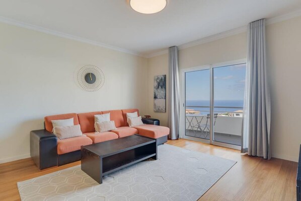 A living room with a sofa, a coffee table, and a view of the sea through a sliding glass door.