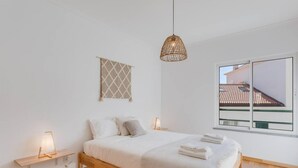 Une chambre à coucher au style minimaliste, avec un lit en bois, des lampes de chevet, un luminaire suspendu en fibres tressées, une grande fenêtre donnant sur un toit recouvert de tuiles et un élément décoratif géométrique sur le mur.