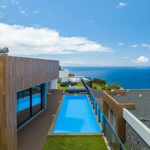 A modern pool area with a clear view of the ocean, a wooden deck, and a glass-enclosed pool. - Casa Mozart I in Estreito da Calheta (Calheta)