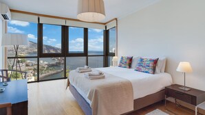 Une chambre d’hôtel moderne avec une grande fenêtre, un lit recouvert d’une courtepointe blanche, des oreillers colorés, une table de chevet avec une lampe, et une vue sur le paysage urbain et l’eau.
