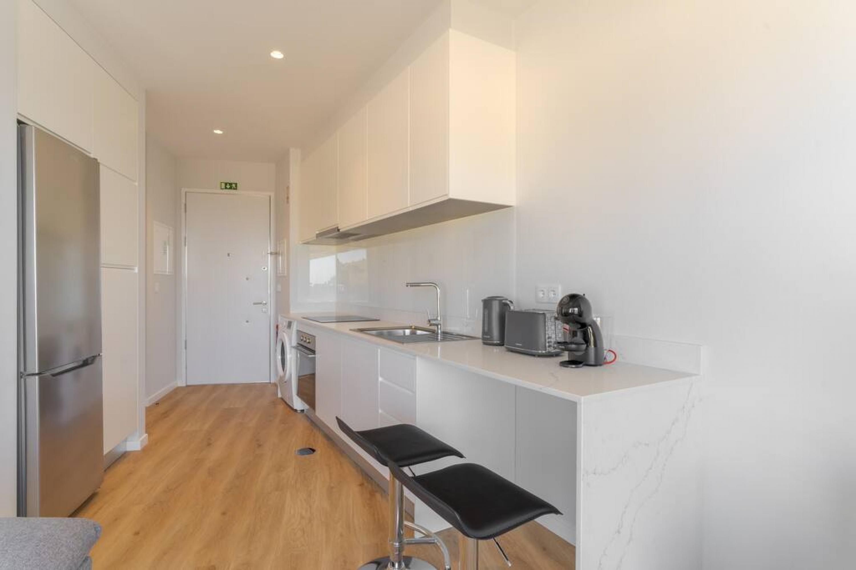 A modern kitchen with white cabinetry, a stainless steel refrigerator, a built-in oven, a sink, and two black bar stools.